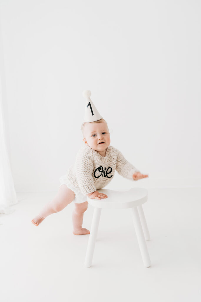 Baby boy in gorgeous ONE birthday jumper standing up on his cake smash photoshoot in Cheshire